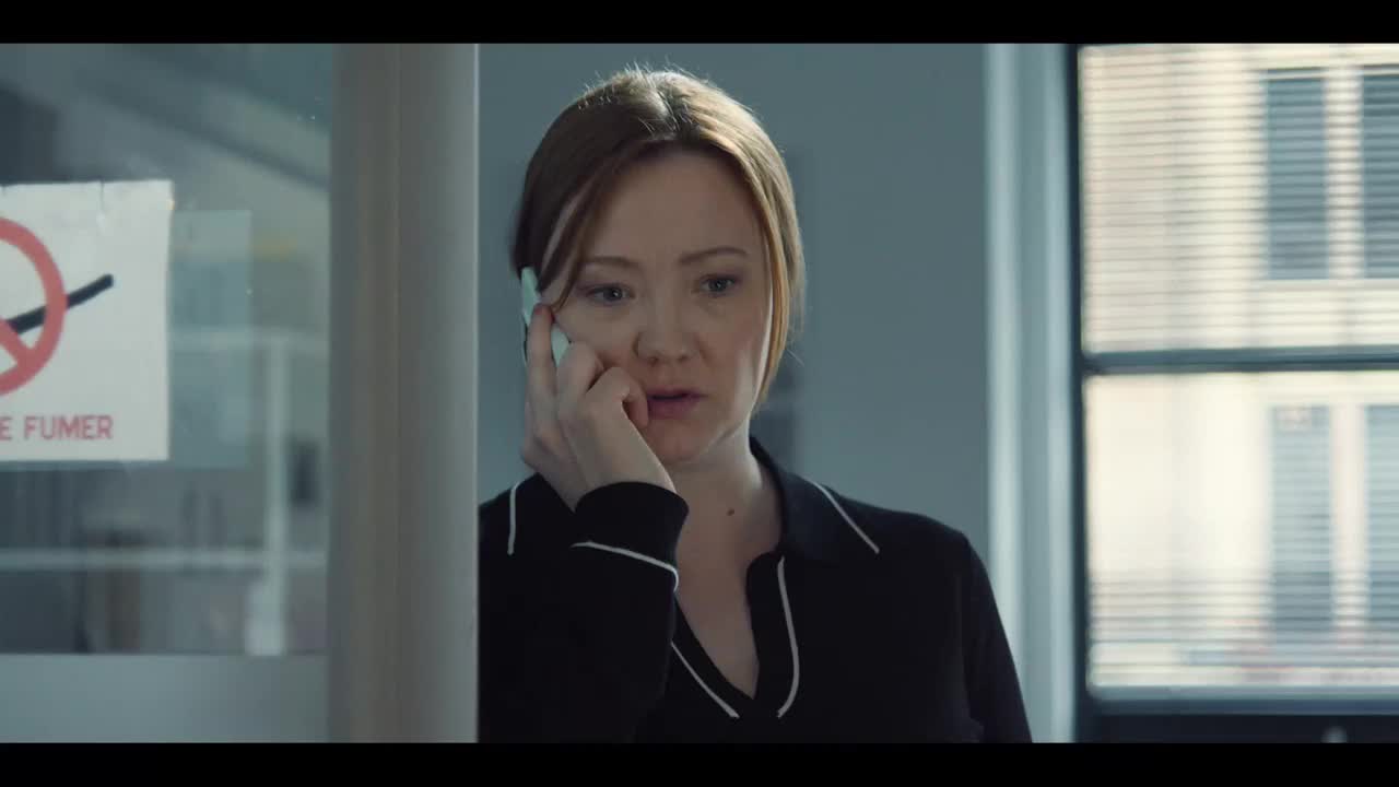 She's holding a phone to her ear, listening intently, her expression a mix of concern and concentration. The no smoking sign on the glass door behind her is ...