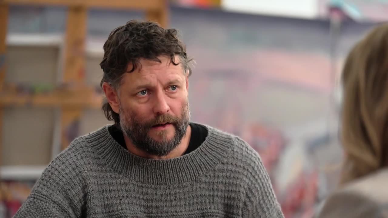 A man with a beard and curly hair, wearing a gray sweater, is looking intently at someone off-camera. Behind him, a colorful painting is visible, suggesting ...