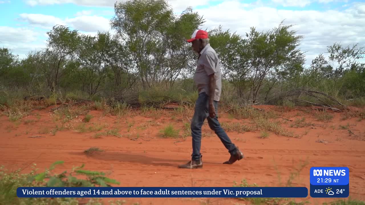 A man in a red and white cap walks along a red dirt track, his boots kicking up a little dust. The Australian bush stretches out behind him, under a partly c...