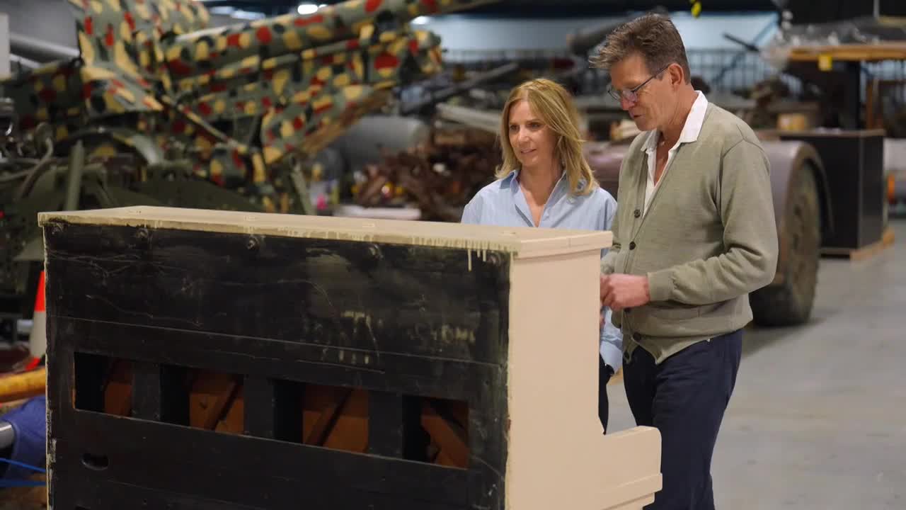A man and a woman are standing beside a piano, examining it closely. The instrument's black front contrasts with its cream-colored sides.