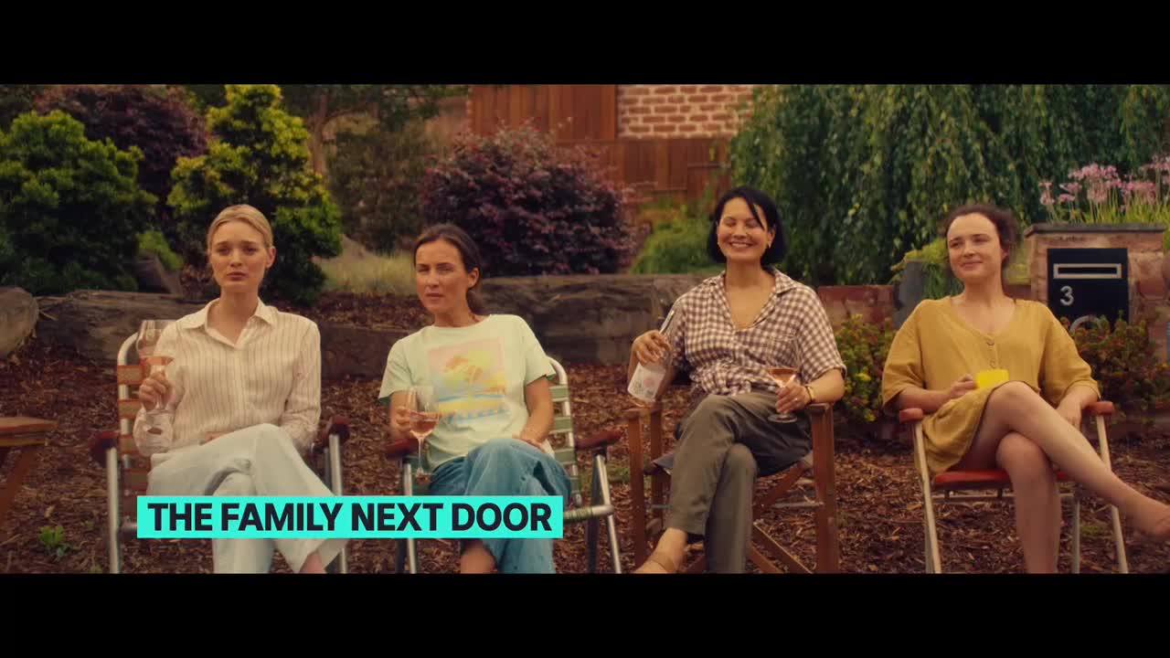 Four women are seated in folding chairs, each holding a glass of wine. They're in a backyard, the house number visible on a mailbox behind them.