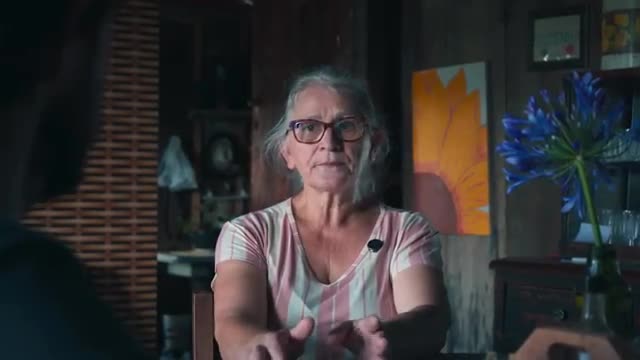 A woman with gray hair and glasses gestures with her hands, speaking to someone off-camera. A small microphone is clipped to her shirt, suggesting a Rede SPT...