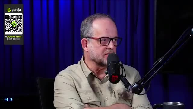 A man with glasses is speaking into a microphone, his face lit by the studio lights. Behind him, a blue curtain forms the backdrop for what looks like a Braz...