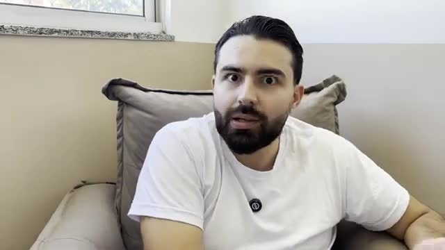 A man with wide eyes and a surprised expression is seated, likely in his home. He's wearing a white t-shirt and looking directly at the camera.
