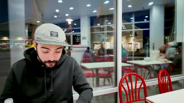 A man in a gray cap and black hoodie is looking down, seemingly focused on something in front of him. Through the window, other people are seated at tables i...