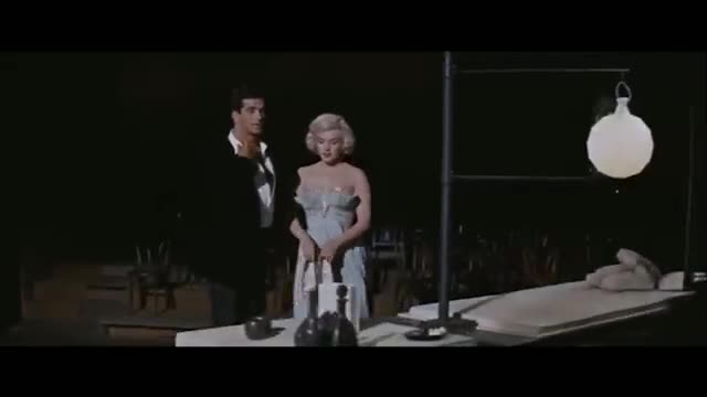 A man in a black suit and a woman in a pale blue dress stand near a table. A white, spherical lamp hangs from a metal frame above a long, flat surface.