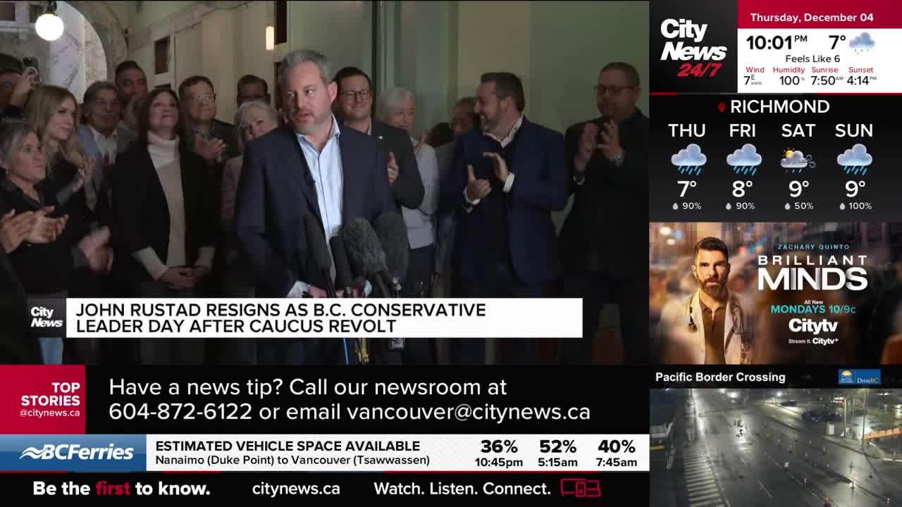 John Rustad, surrounded by a crowd, stands before microphones, speaking to the press. Behind him, people clap and watch as the CityNews Vancouver headline an...