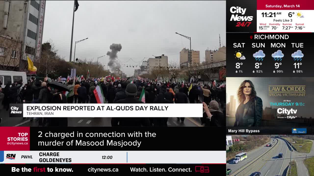 A thick plume of smoke rises into the sky above a crowded street filled with people holding flags. CityNews Vancouver reports an explosion occurred at the Al...