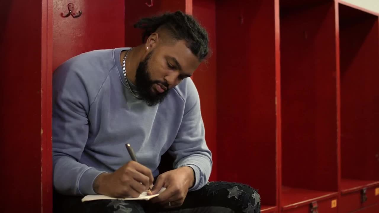 A man sits in a locker room, his pen moving across a small notepad. The deep red of the lockers behind him contrasts with his light blue sweatshirt.
