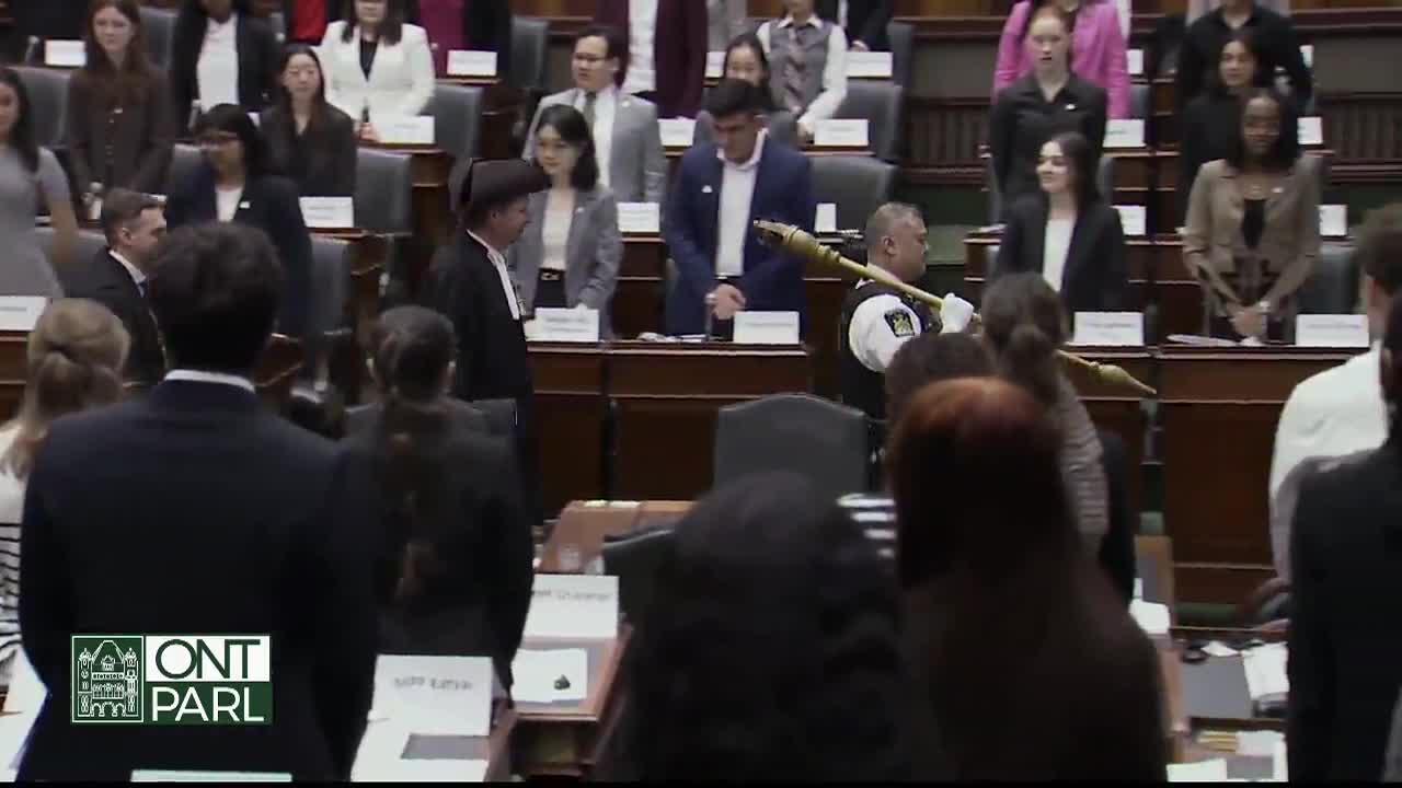 In the Legislative Assembly of Ontario, a man in a uniform carries a ceremonial mace through the room. The Speaker, in traditional robes, is also present as ...