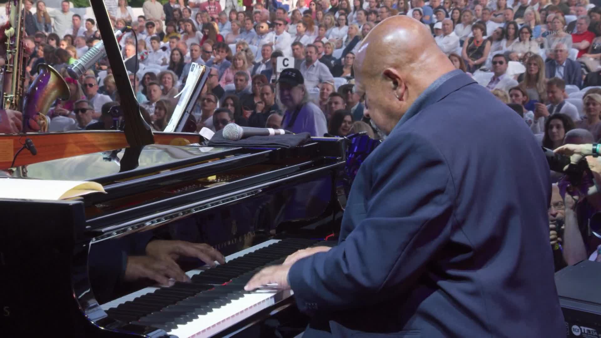 A man in a blue suit is hunched over a grand piano, his fingers dancing across the keys. The audience, packed tightly in the background, watches intently as ...