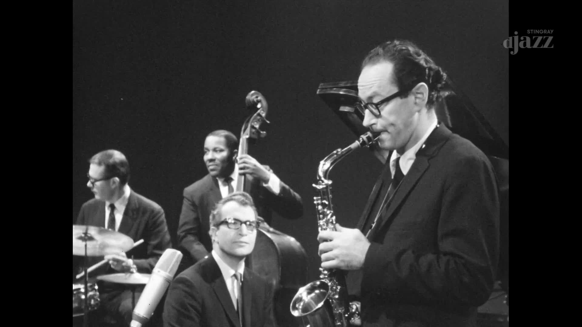 The saxophonist, in a dark suit and glasses, blows into his instrument, his cheeks puffed out. A bassist and drummer provide the rhythm, while a pianist sits...