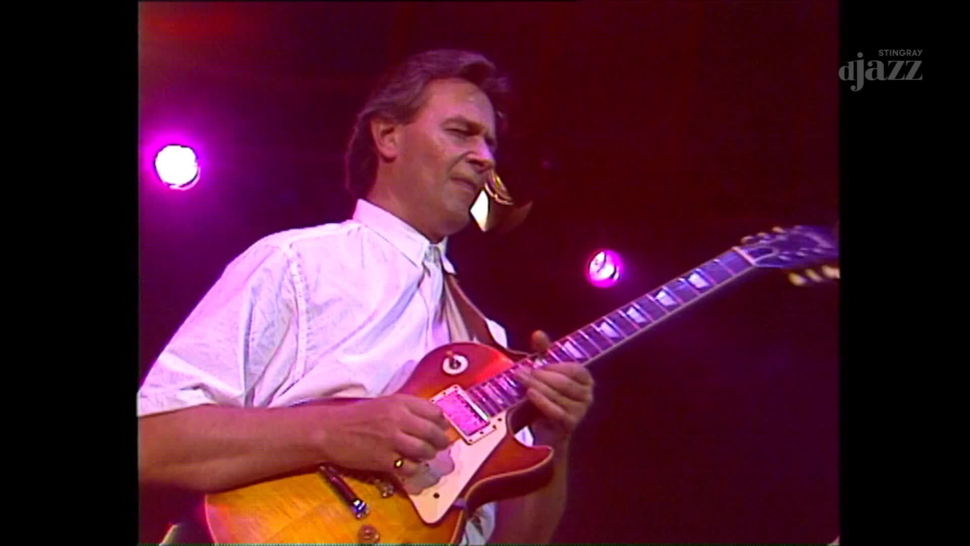 The guitarist, bathed in purple light, is focused on the fretboard, his fingers dancing across the strings. He's playing a classic-looking electric guitar, a...