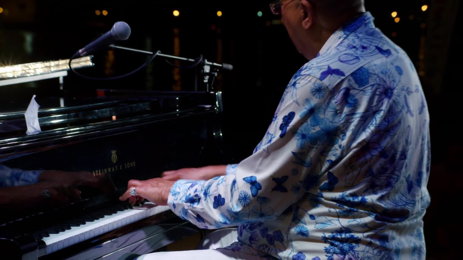 The musician's hands dance across the piano keys, the notes echoing in the dim space. His patterned shirt, a splash of blue and white, contrasts with the bla...