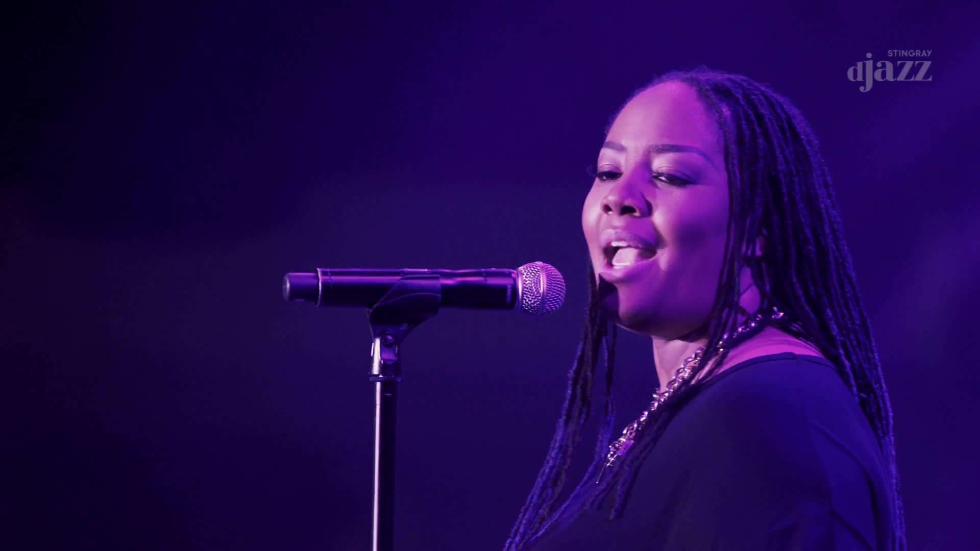 A woman with long braids sings into a microphone, her mouth open in mid-note. The Stingray DJAZZ logo is visible in the corner, and the stage is bathed in pu...