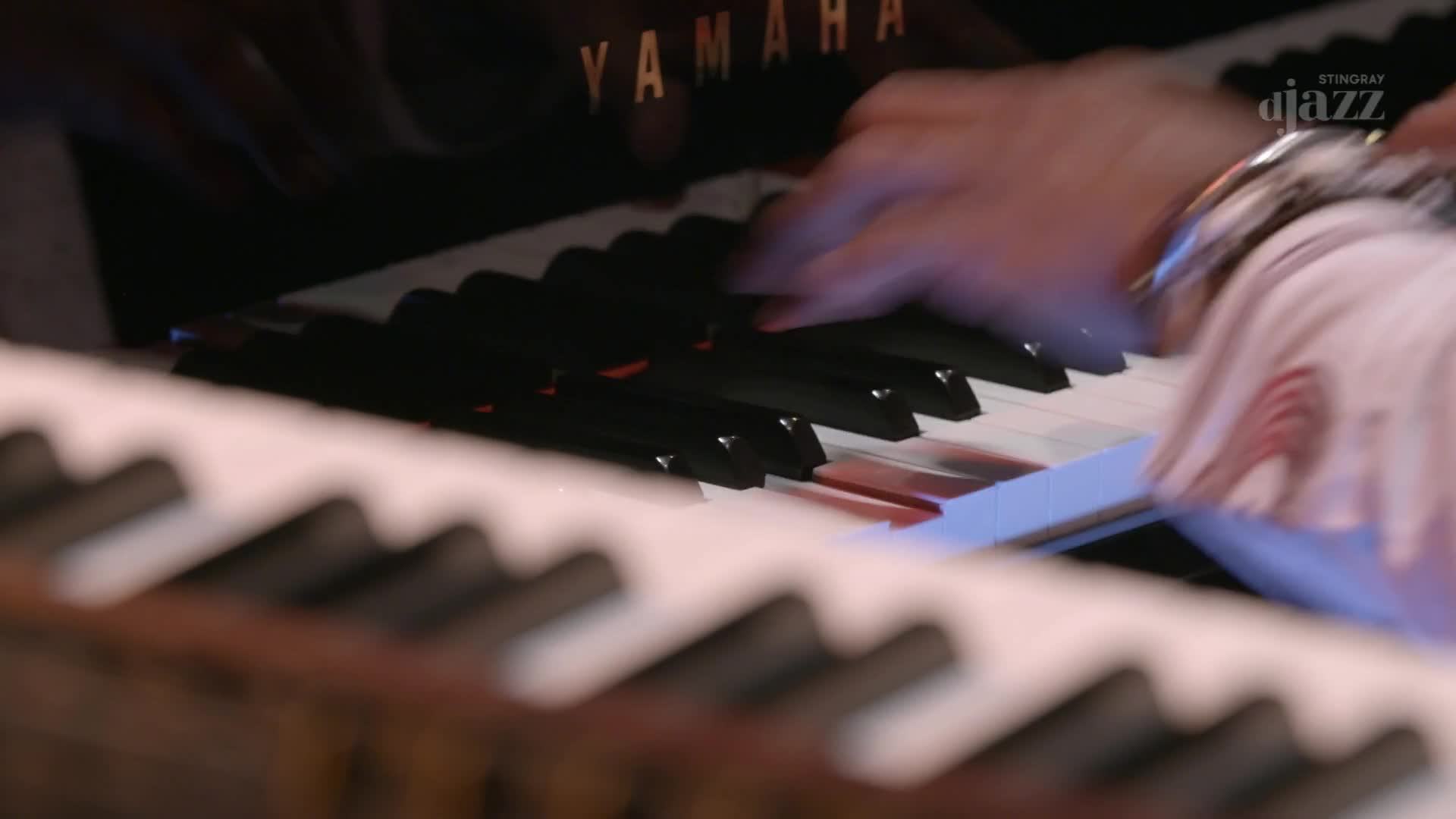 Fingers dance across the piano keys, a Yamaha logo visible above. The musician's wrist, adorned with a silver bracelet, is in motion, close to the Stingray D...