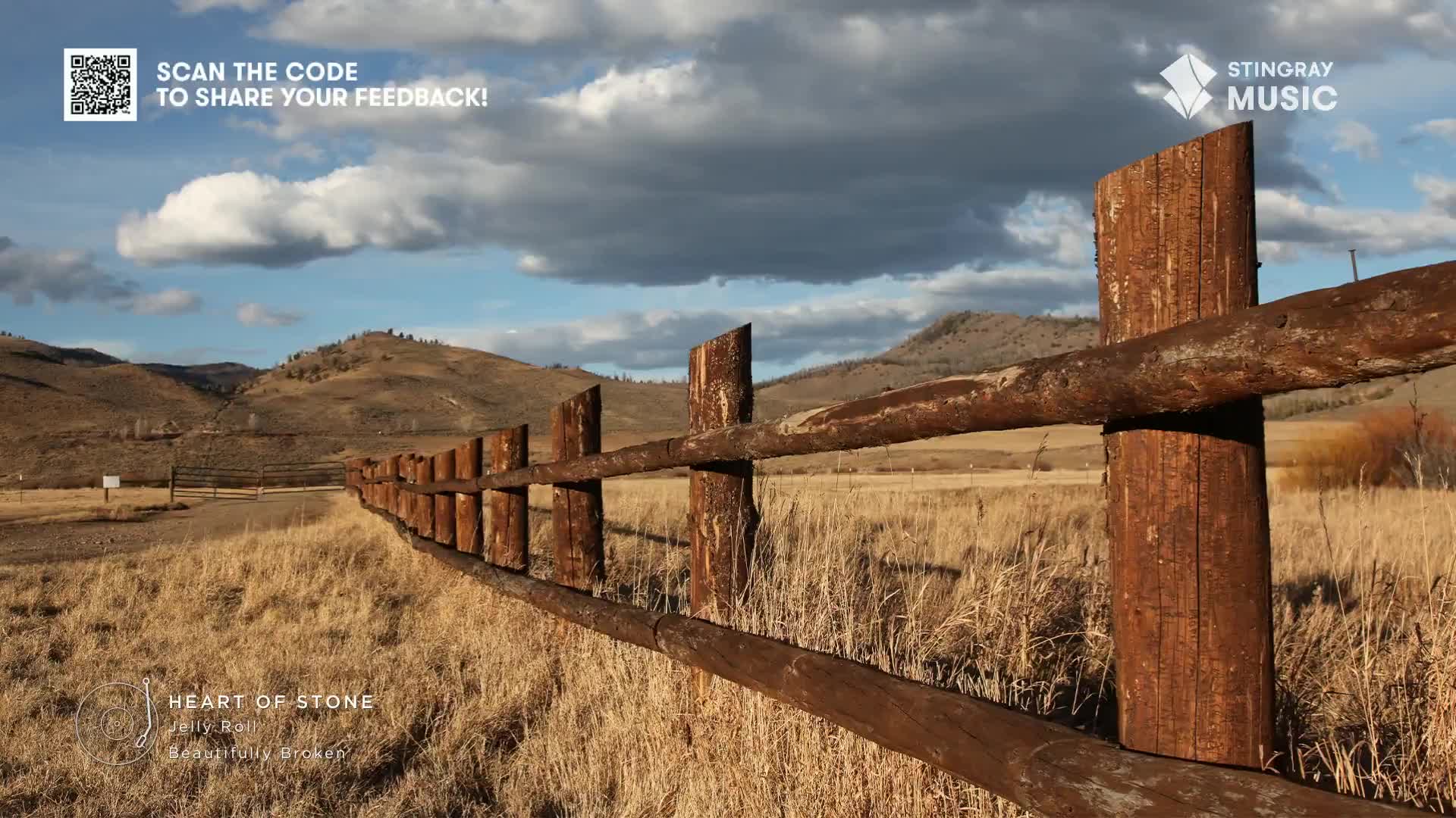 A weathered wooden fence stretches across a field of dry grass, leading the eye towards the distant hills. The sky is a mix of blue and cloud, and the air fe...
