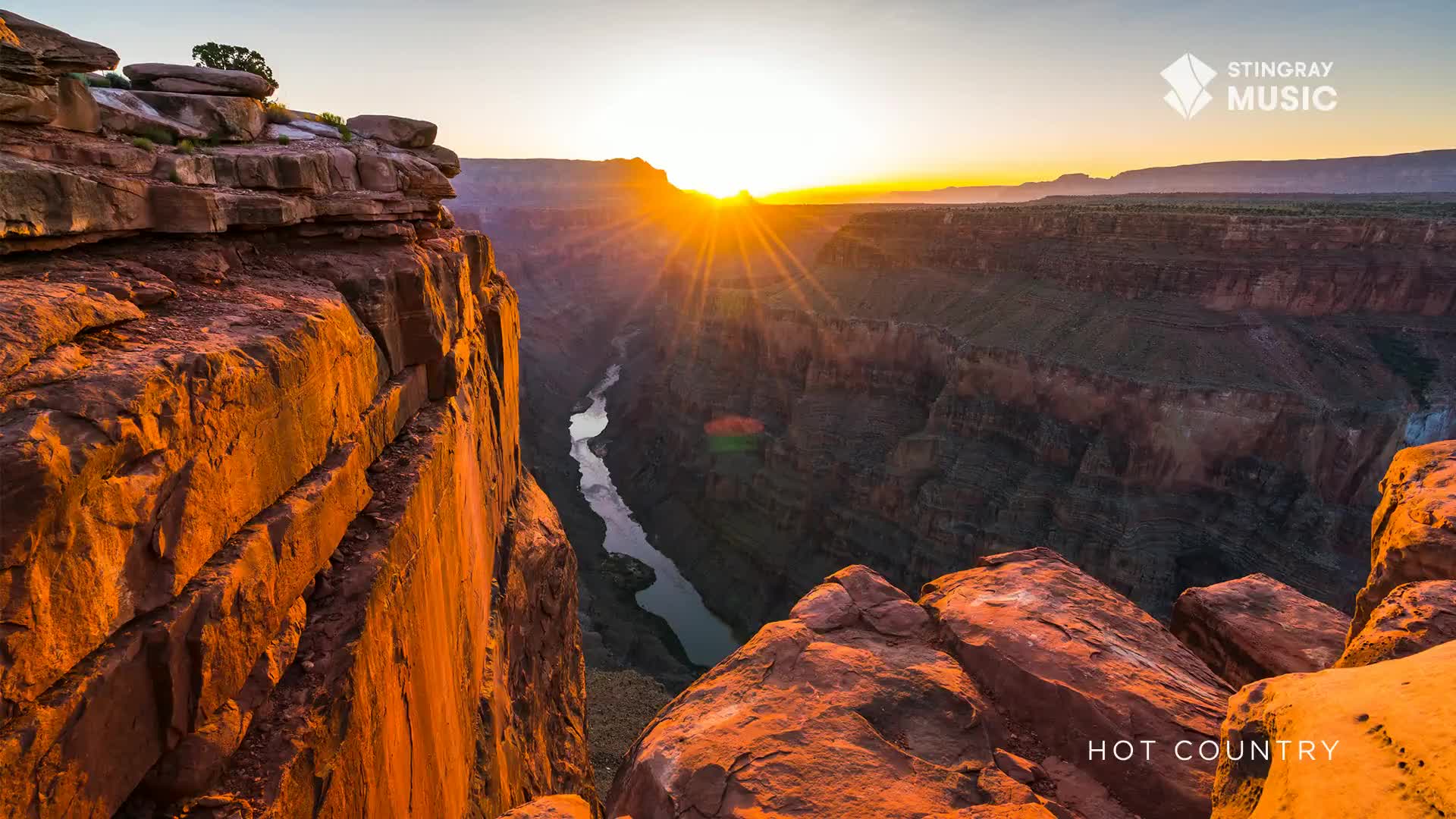 The sun bursts over the canyon rim, its rays painting the rock faces in warm hues. A river snakes through the deep valley below, reflecting the golden light.