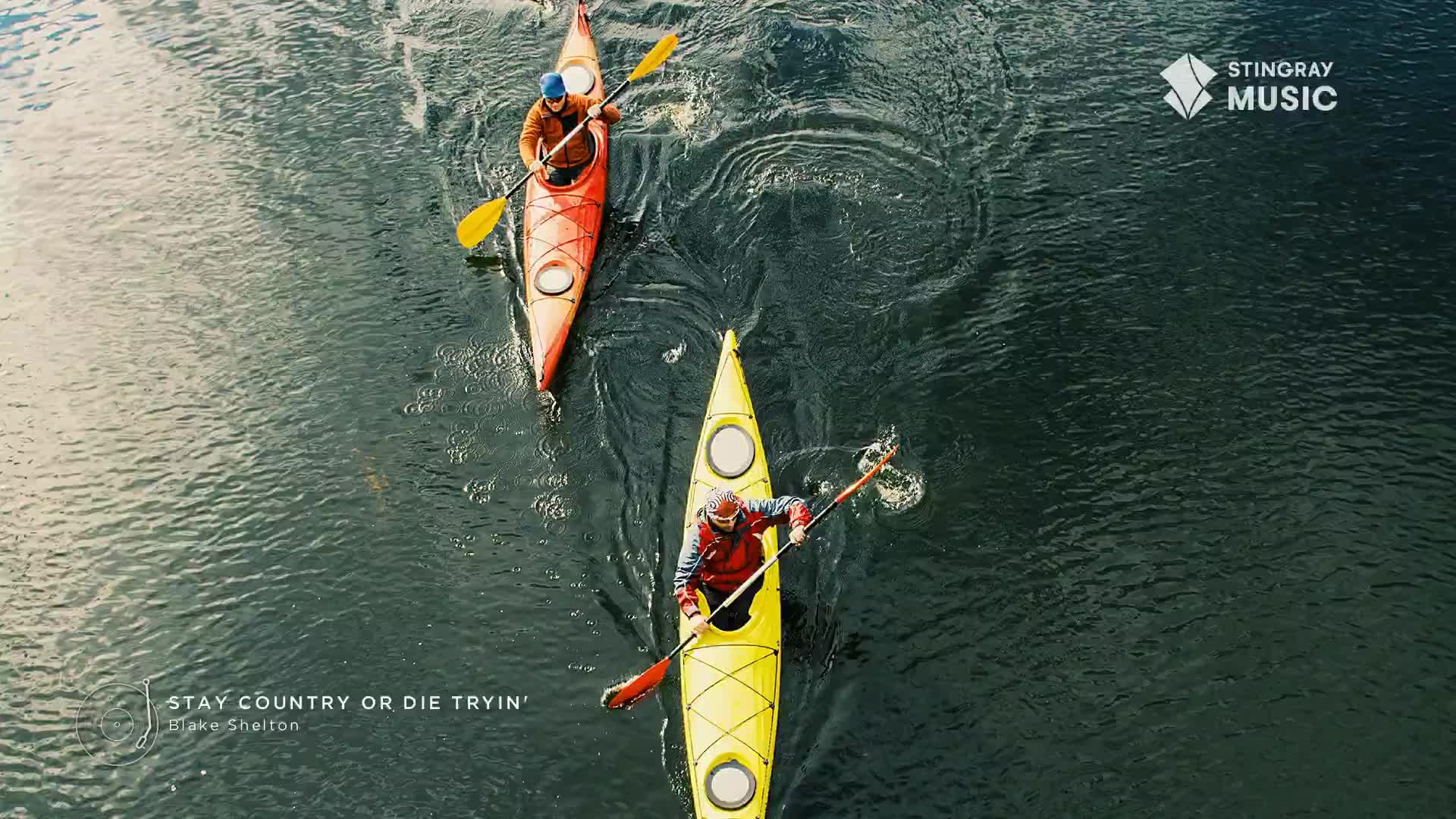 Two kayakers, one in a yellow boat and the other in red, paddle across the dark water, leaving swirling trails behind them. The Stingray Hot Country logo sit...