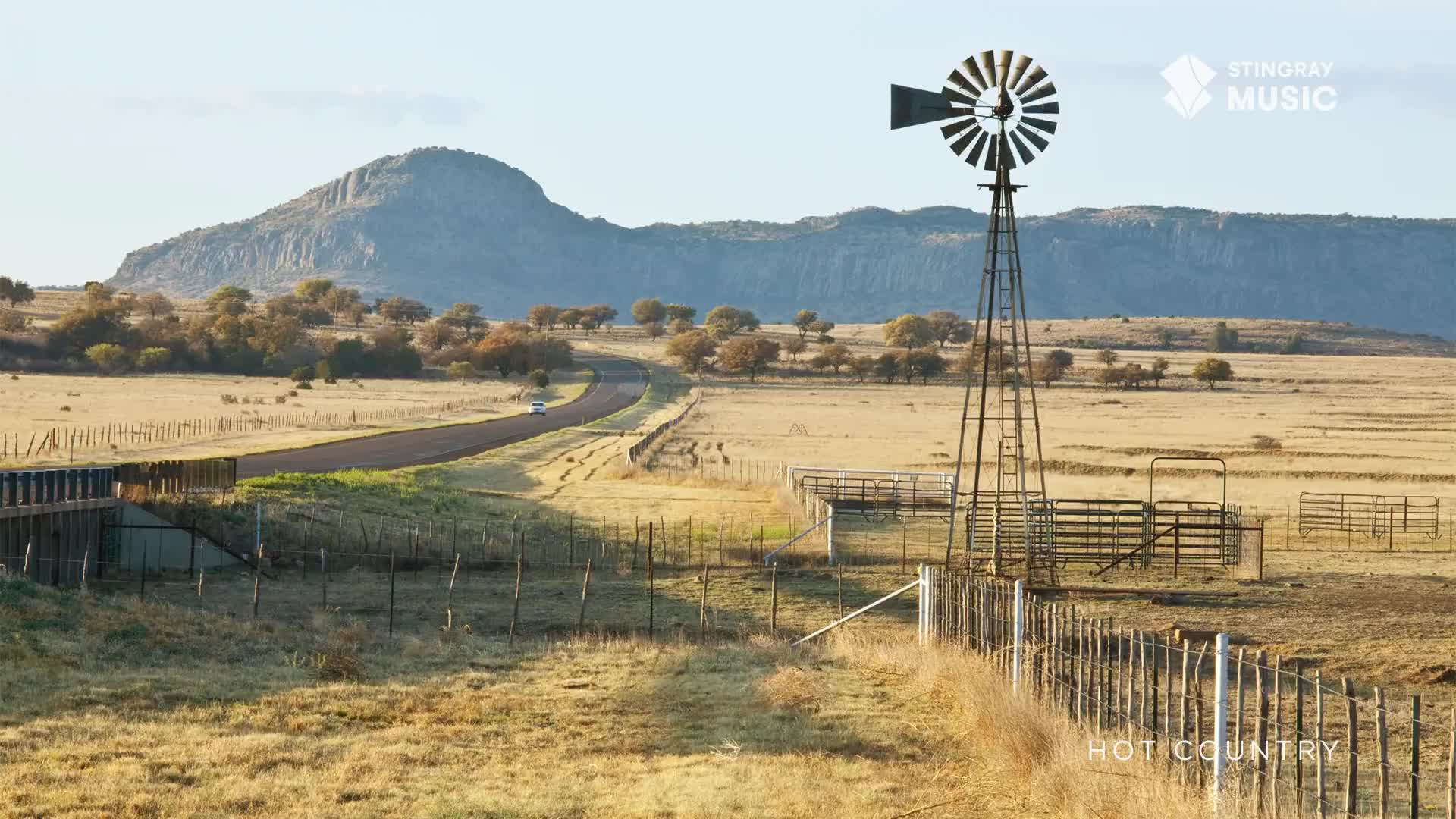 A lone car travels along a winding road, disappearing into the distance. The blades of a windmill stand still against the backdrop of a large, rocky hill.