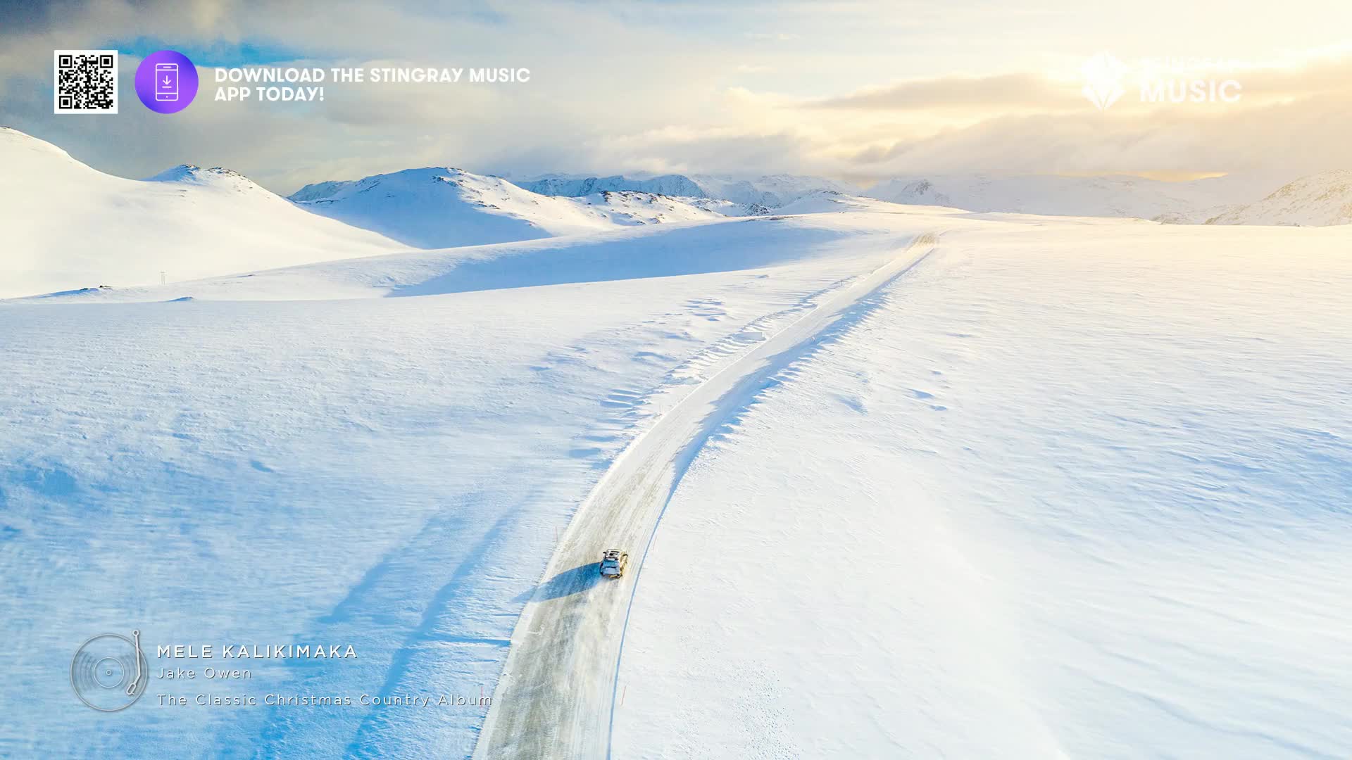 A car, small against the snowy landscape, drives along a road cut through the mountains. The sky is bright with a hint of sun, and the whole scene feels cold.