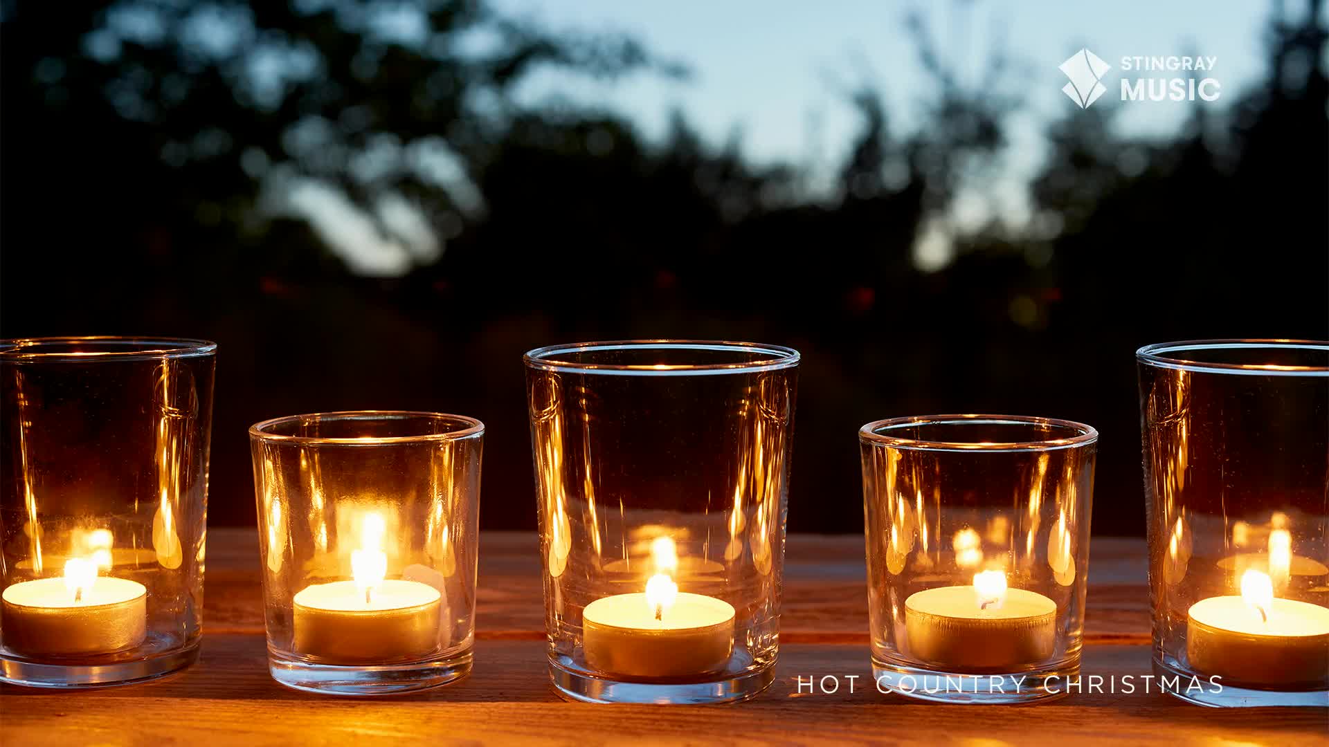 Five small candles flicker inside clear glass holders, casting a warm glow on the wooden surface. The scene is set outdoors, under a dark sky, and the Stingr...