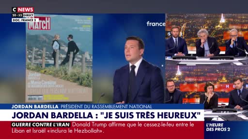 A man in a suit stands at a podium, speaking into a microphone. To his left, a magazine cover shows two figures on a hillside. To his right, a panel of comme...