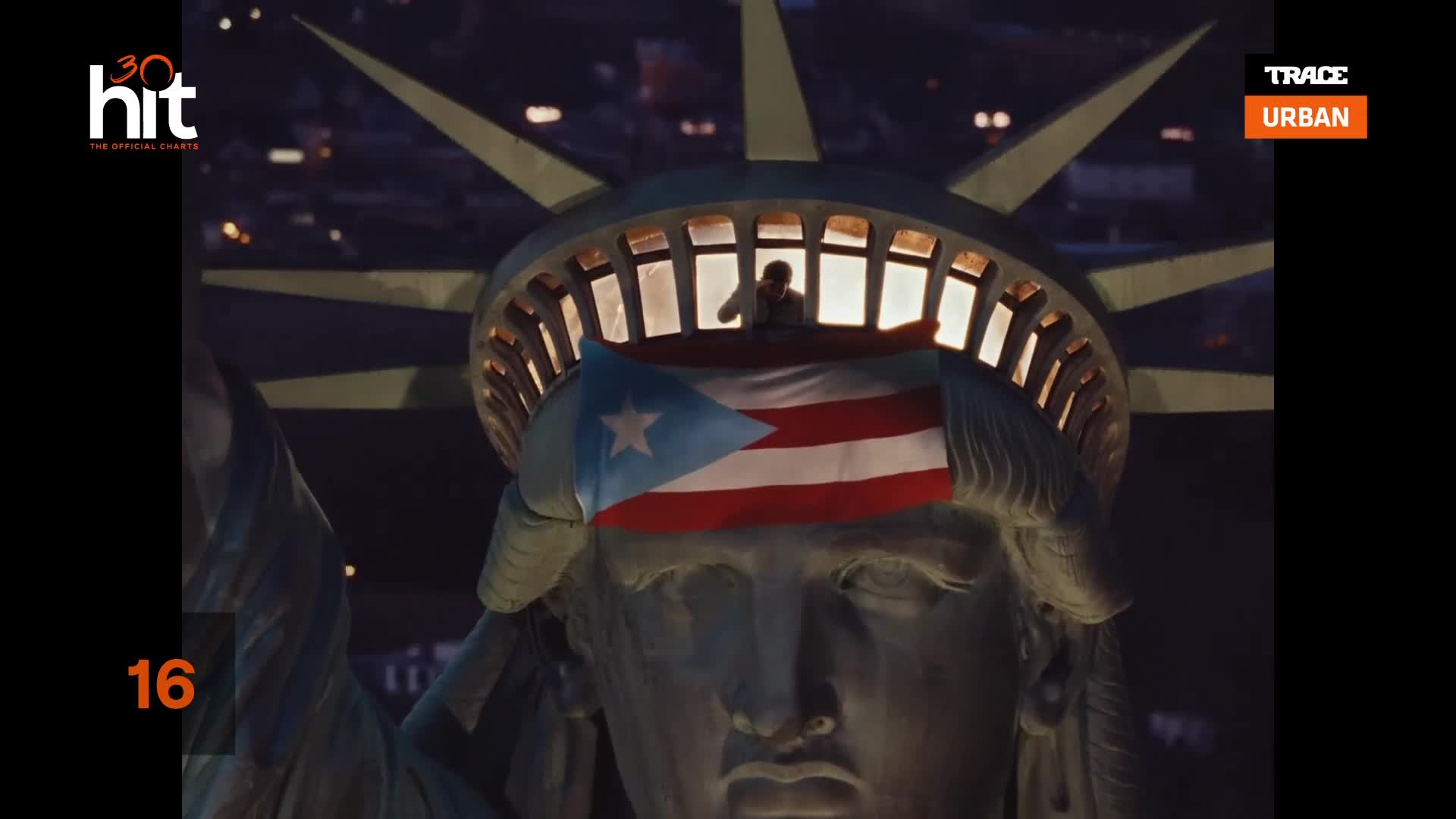 The Statue of Liberty's crown is lit from within, and a Puerto Rican flag is draped across her forehead. A person stands in the crown's opening, silhouetted ...