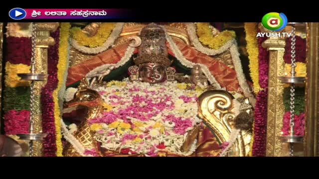 A decorated deity is the focus, covered in flowers and adorned with a jeweled crown. The scene is framed by ornate gold pillars and broadcast on Ayush TV.