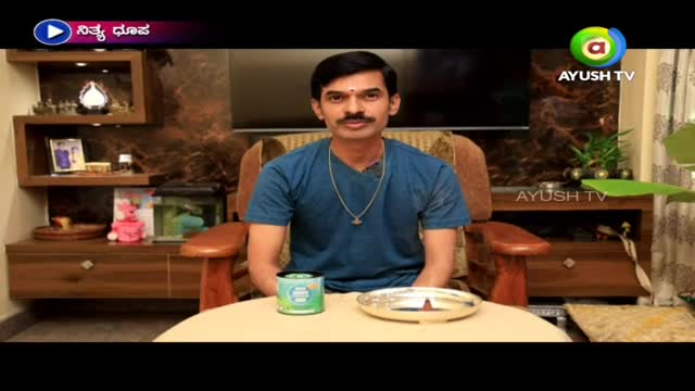 A man in a blue shirt sits in a wooden chair, facing the camera with a calm expression. A silver plate and a green tin sit on the table in front of him, whil...