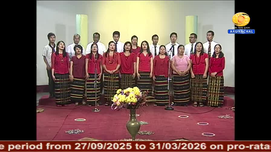 A choir, dressed in red tops and patterned skirts, stands on a red carpet, ready to sing. Behind them, men in white shirts and ties stand, likely part of the...