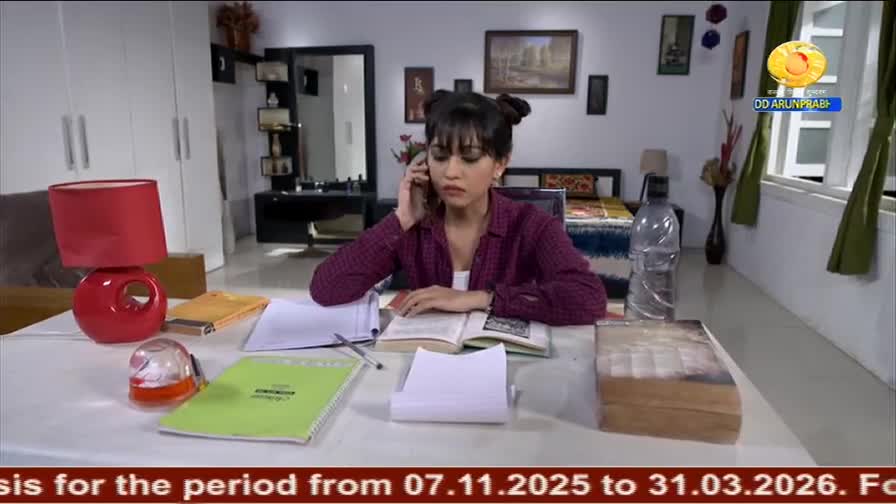 A young woman in a maroon shirt is talking on the phone while studying at a table, books and papers scattered around. The DD Arun Prabha logo is visible in t...