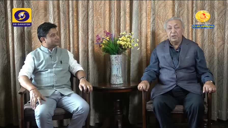 Two men sit facing each other on a set, likely for a DD Arun Prabha interview. The man on the right, in a navy blue jacket, appears to be speaking.