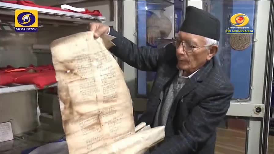 An older man, wearing a black cap, carefully holds up a large, aged document, its surface covered in faded script. The DD Arun Prabha logo is visible as he d...