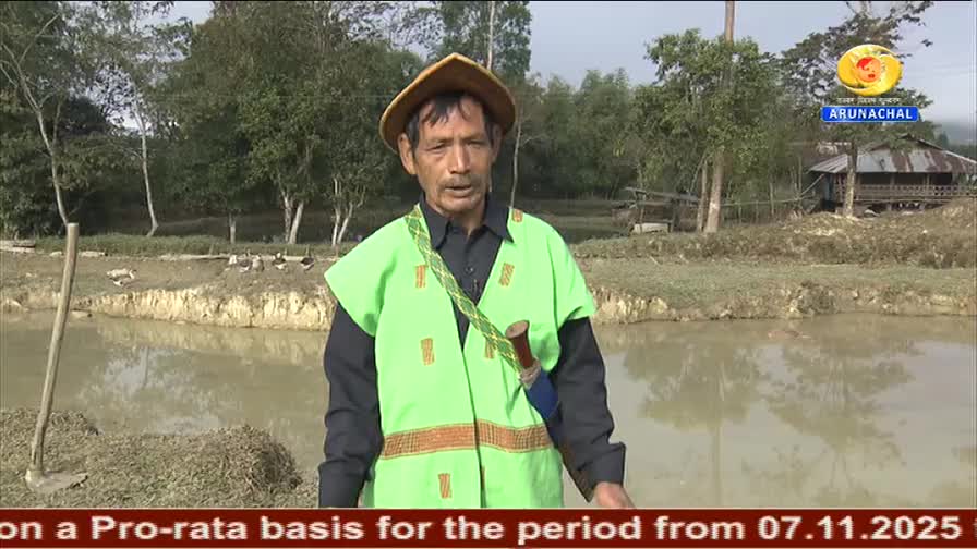 The man, wearing a green vest and hat, is speaking near a body of water. Behind him, a DD Arun Prabha logo is visible, and a caption scrolls across the botto...
