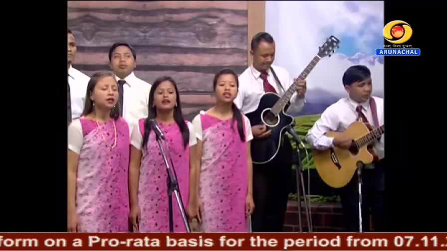 On the DD Arun Prabha set, a group of singers in pink patterned dresses and white blouses stand in a row, harmonizing into microphones. Two guitarists accomp...