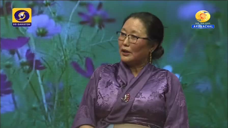 A woman in a purple robe speaks, framed by a floral backdrop, with the DD Gangtok and Arunachal Pradesh logos visible. She gestures with her hands as she spe...