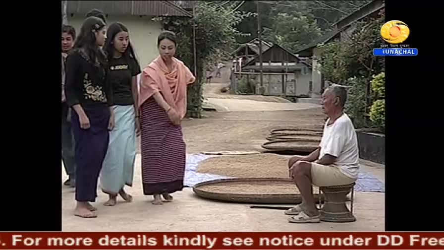 A group of women in traditional dress stand facing an older man seated on a stool, with drying grains spread out on large, round trays nearby. The scene appe...