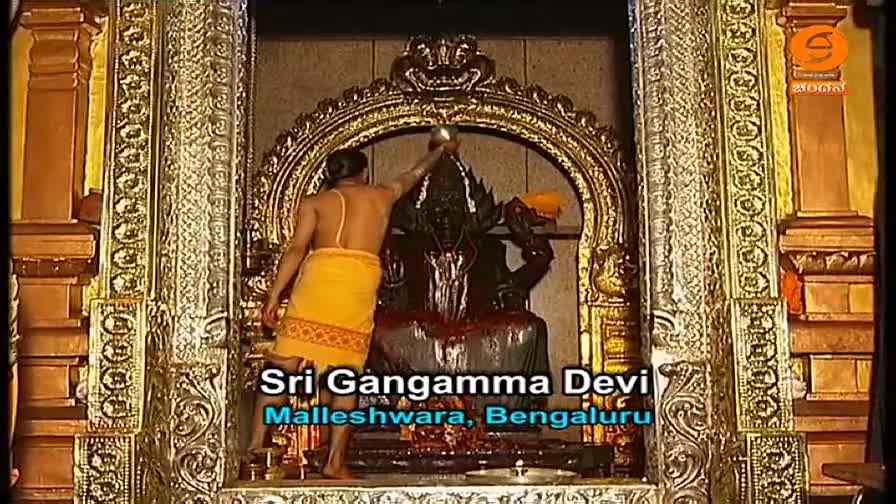 A man in a yellow dhoti pours liquid from a small vessel over a dark statue of Sri Gangamma Devi. The scene is being broadcast on DD Chandana, with text iden...