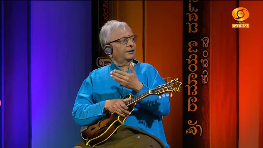 A man in a blue kurta gestures with his right hand while holding a mandolin. He's wearing glasses and a headset, likely on the DD Chandana set in India.