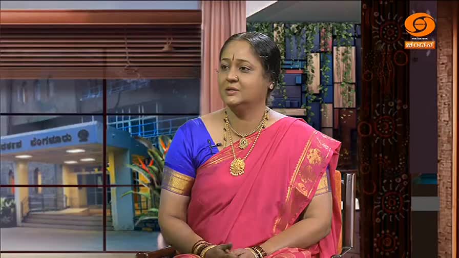 A woman in a pink sari and gold jewelry sits and speaks, framed by a window showing a building. The DD Chandana logo is visible in the corner.