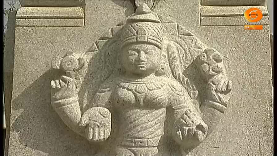 A stone carving of a figure, with a serene face and ornate headdress, is etched into a wall. The figure's arms are outstretched, and the hands hold objects t...