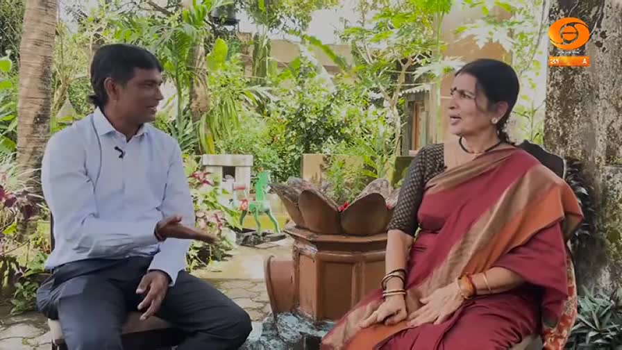 A man in a light blue shirt gestures as he speaks, while a woman in a red sari listens intently. The scene, set outdoors, is likely a DD Chandana interview.