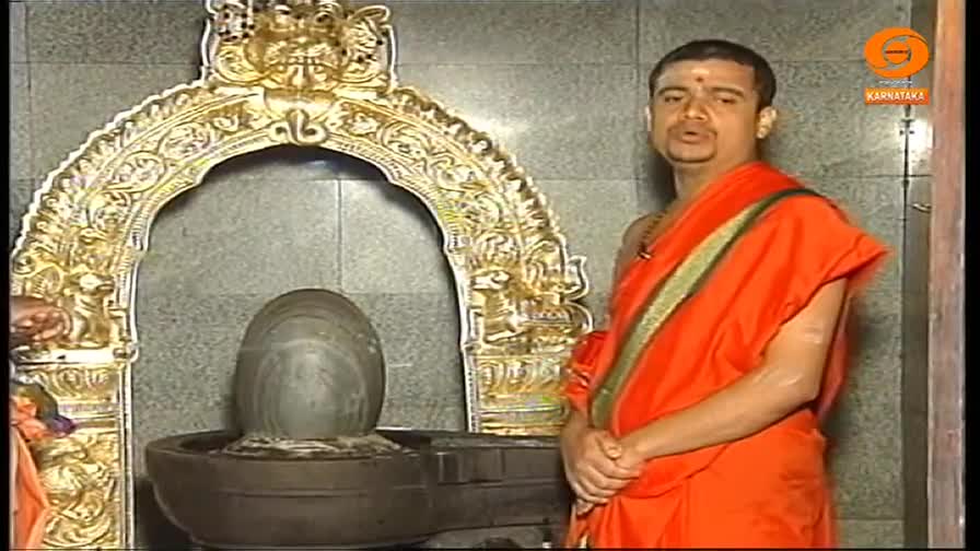 A man in orange robes stands before a shrine, speaking directly to the camera. Behind him, a stone lingam sits in a basin, framed by an ornate golden arch.