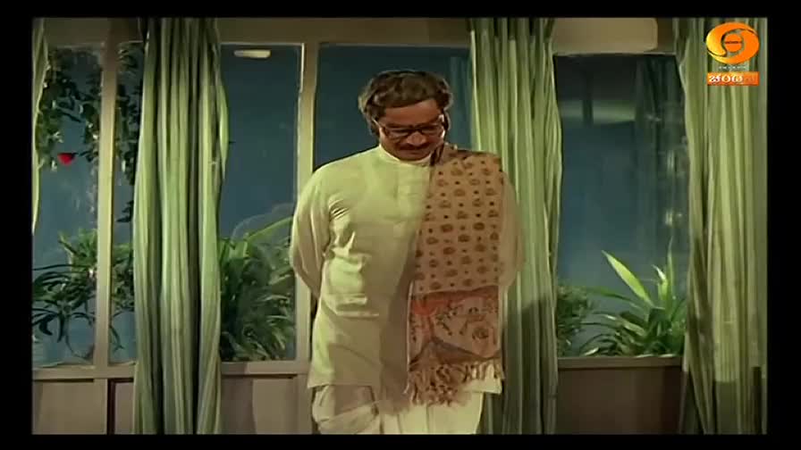 A man in a kurta and shawl stands in a room, his gaze fixed downwards. Behind him, the window reveals a glimpse of greenery and the logo of DD Chandana.