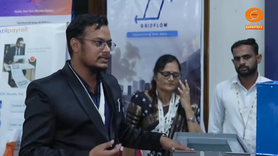 A man in a blazer gestures toward a device with a clear top, while two others look on. The backdrop includes a sign for "Gridflow" and a logo for Karnataka.