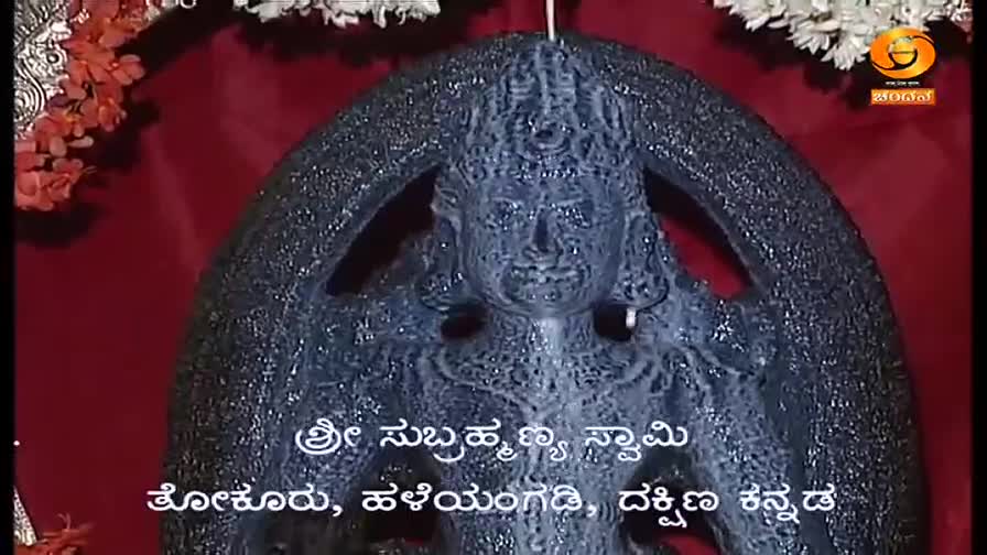 A dark, textured sculpture of a deity is centered, framed by a red backdrop and floral accents. Below, text in Kannada identifies the location as Sri Subrahm...