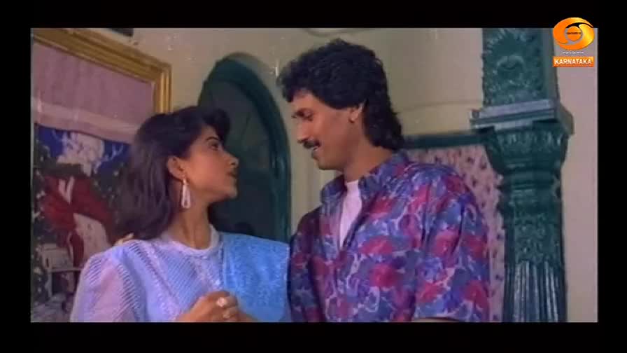 A man in a patterned shirt gazes at a woman wearing a light blue top, both of them in a scene from what looks like a DD Chandana production. They appear to b...