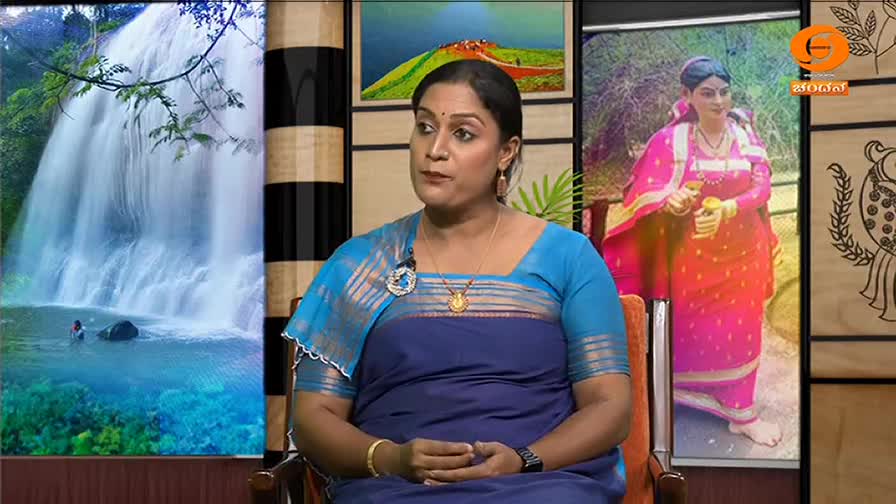 DD Chandana, dressed in a blue saree, sits in a studio set, speaking to someone off-screen. Behind her, a waterfall and a portrait of a woman in traditional ...