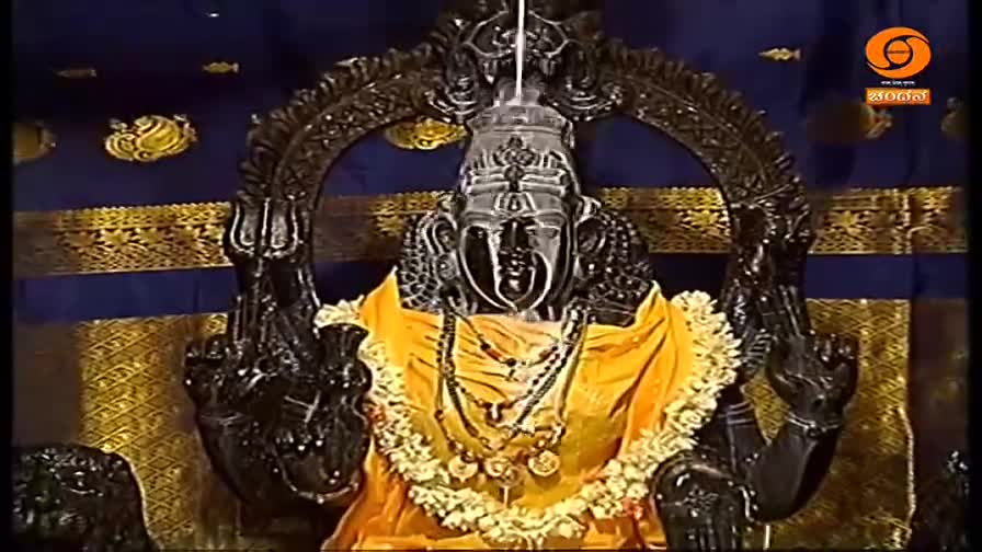 A dark, ornate statue of a deity is centered, draped in a yellow cloth and garlands. The statue is framed by an elaborate arch against a backdrop of blue and...